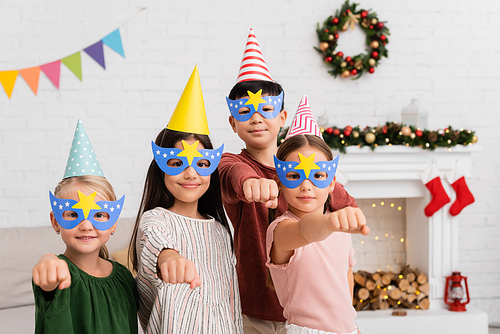 Multiethnic kids in party masks and caps gesturing at camera at home