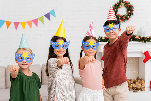 Cheerful interracial kids in party caps and masks gesturing during birthday party at home