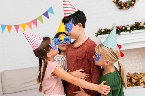 Smiling kids hugging asian friend in party cap and mask during birthday celebration at home