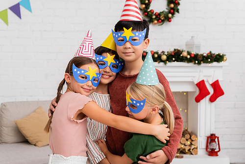 Cheerful interracial children in party caps and masks hugging during birthday celebration at home