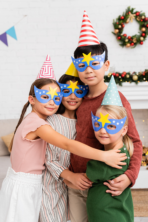 Positive interracial kids in party caps and masks hugging and looking at camera at home