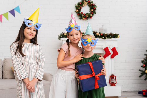 Smiling girls in party caps and masks hugging friend with present near christmas decor at home