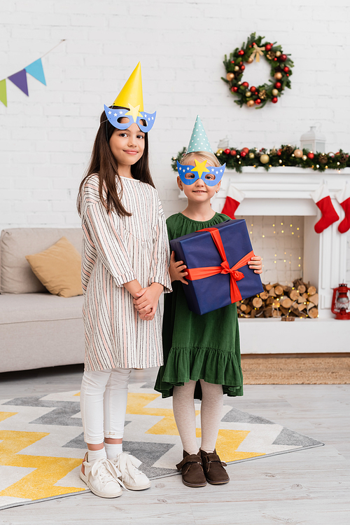 Smiling girls in party caps and masks holding present during birthday celebration in living room
