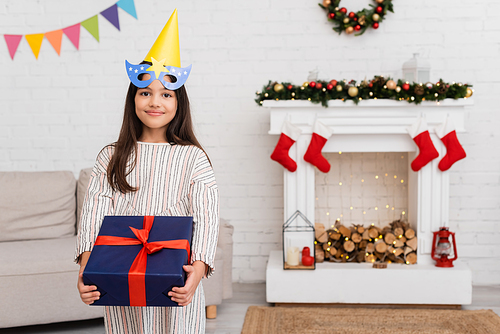Smiling girl in party mask and cap holding gift with ribbon during birthday celebration in winter at home
