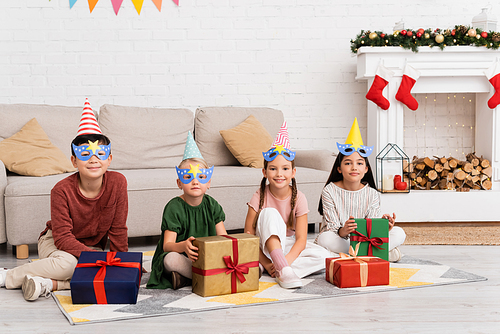 Smiling multiethnic kids in party caps and masks looking at camera near presents during birthday at home