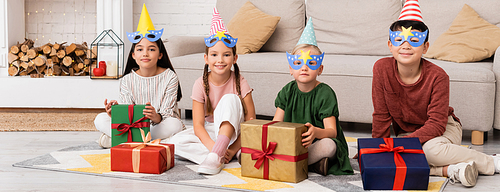 Positive multiethnic children in party caps and masks looking at camera near presents at home, banner