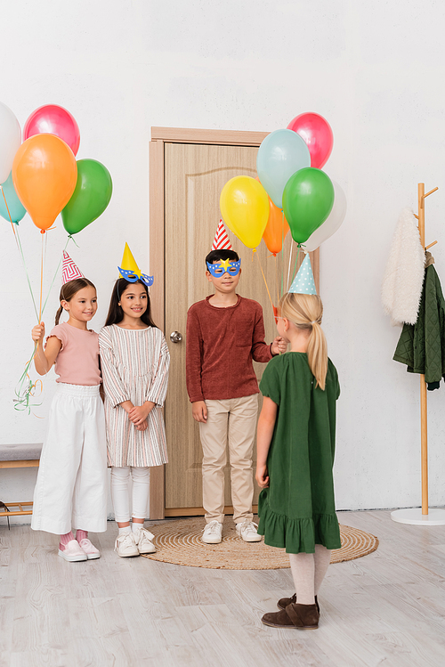 Interracial kids in party masks and caps holding balloons near friend in hallway
