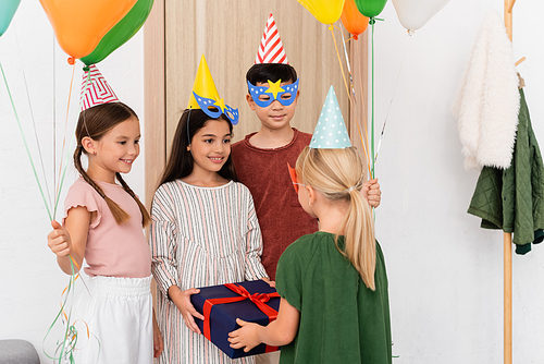 Cheerful interracial children holding gift and balloons near friend in hallway at home