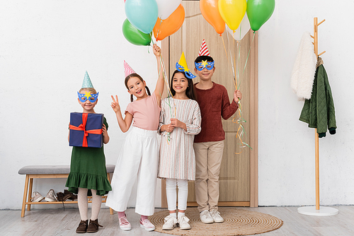 Cheerful multiethnic kids in party caps holding balloons and gift while looking at camera in hallway at home