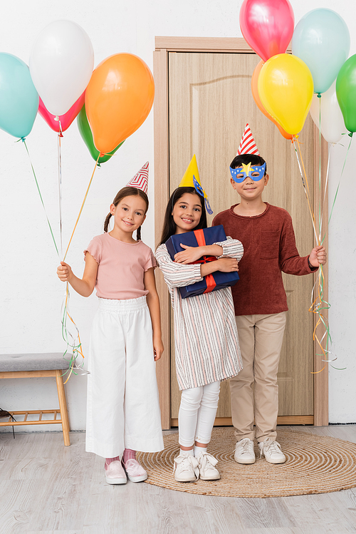 Smiling girl in party cap holding gift box near interracial friends with balloons in hallway at home