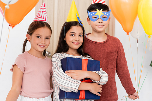Smiling interracial kids in party caps holding gift and balloons in hallway at home