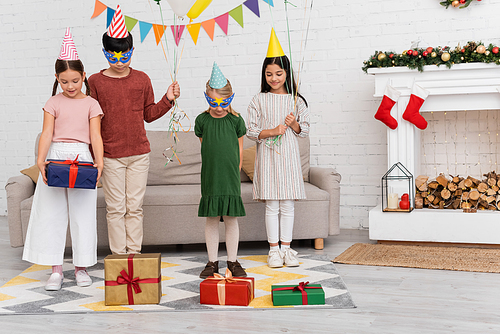 Smiling multiethnic kids in party caps holding balloons near presents and christmas decor at home