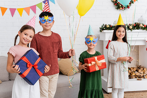 Multiethnic children in party caps holding balloons and presents during birthday celebration in winter at home