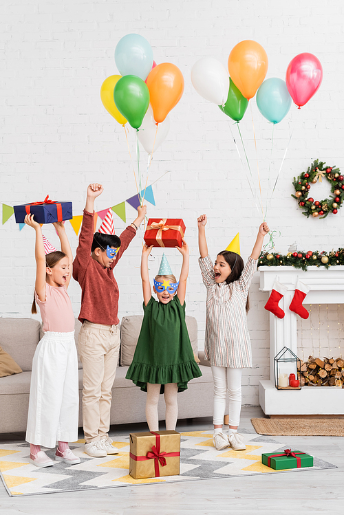 Excited multiethnic children in party caps and masks holding balloons near friend with birthday gift and christmas decor at home