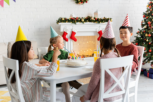 Interracial kids in party caps sitting near birthday cake and orange juice at home in winter