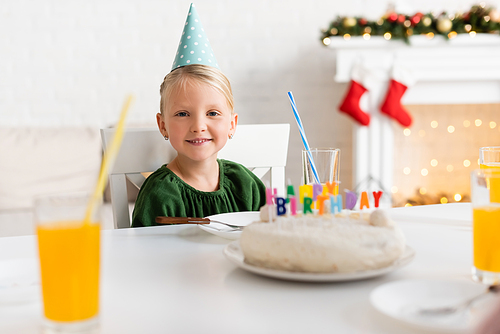 Smiling girl in party cap looking at camera near birthday cake and orange juice at home