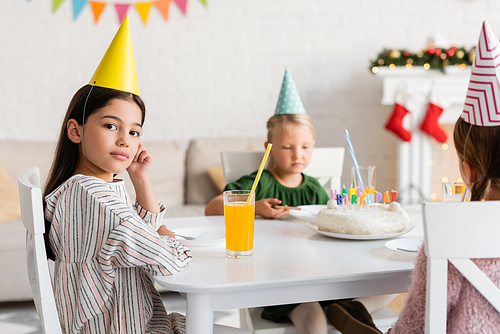 Girl in party cap looking at camera near friends and birthday cake at home