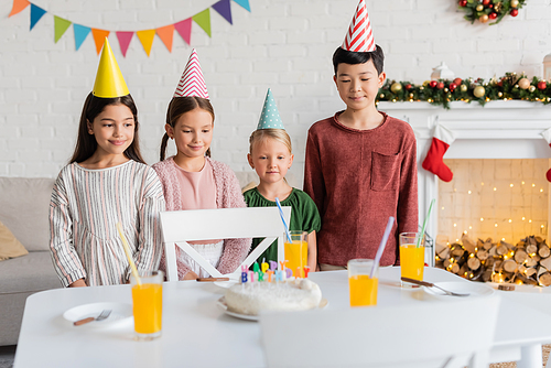 Smiling interracial kids in party caps looking at birthday cake during party at home in winter