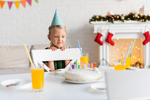 Kid in party cap looking at glasses with orange juice and birthday cake at home