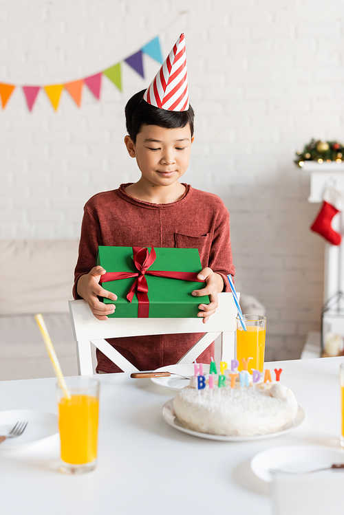 Asian boy in party cap holding gift near birthday cake and orange juice at home