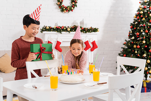 Smiling asian boy in party cap holding present near friend and birthday cake at home in winter
