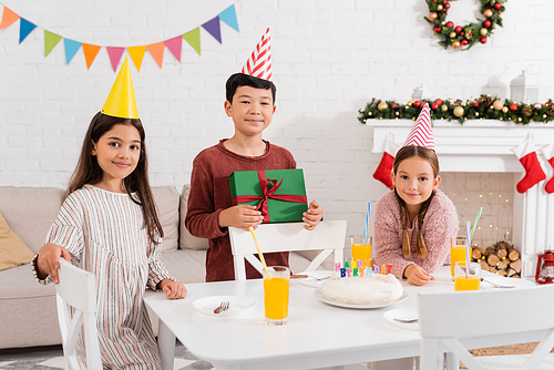 Interracial friends in party caps holding gift near birthday cake at home in winter