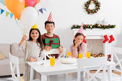 Multiethnic children holding balloons and present near birthday cake at home in winter