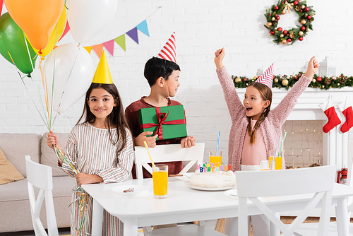 Smiling multiethnic kids in party caps holding balloons and gift near excited friend and birthday cake at home in winter