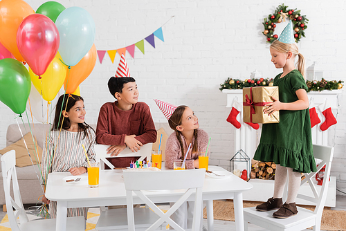 Interracial kids in party caps holding balloons near friend with gift and birthday cake at home
