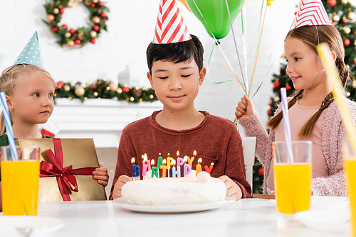 Asian boy in party cap looking at birthday cake with candles near friends with gift and balloons at home