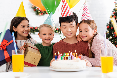 Multiethnic kids in party caps holding presents near birthday cake and orange juice at home