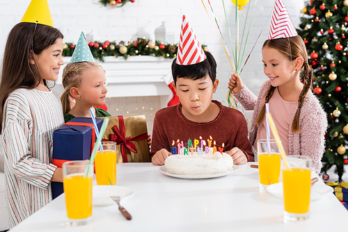 Asian boy in party cap blowing out candles on birthday cake near friends with present at home