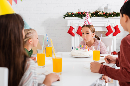 Girl in party cap blowing out candles on birthday cake near friends and orange juice at home