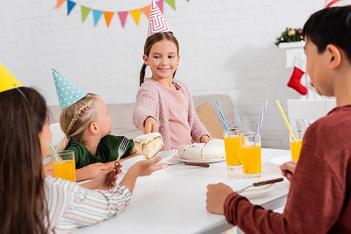 Cheerful child in party cap giving birthday cake to friend during celebration at home