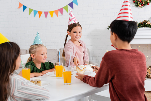 Kid in party cap giving birthday cake to blurred friend near children at home