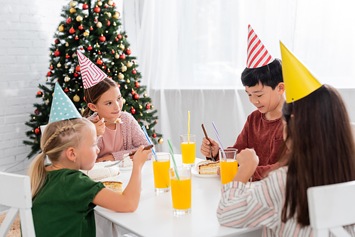 Multiethnic kids in party caps celebrating birthday near cake and orange juice at home