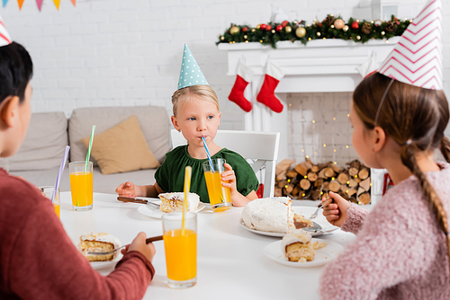 Kid in party cap drinking orange juice near blurred friends and birthday cake at home in winter