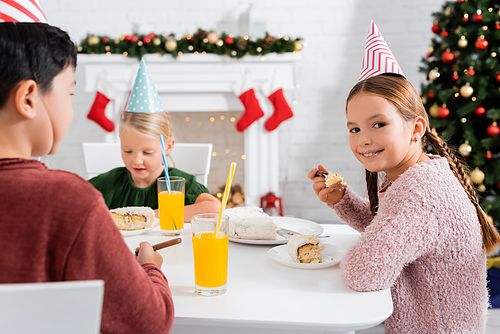 Smiling girl in party cap looking at camera and holding birthday cake near multiethnic friends and orange juice at home in winter