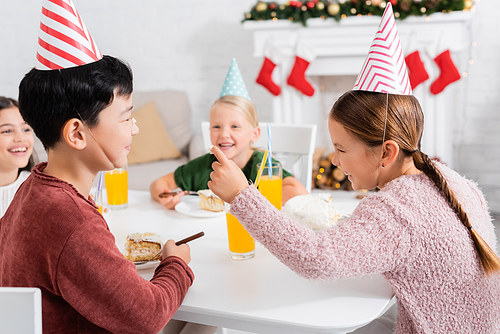 Cheerful kid smearing nose of asian friend with cream of birthday cake at home