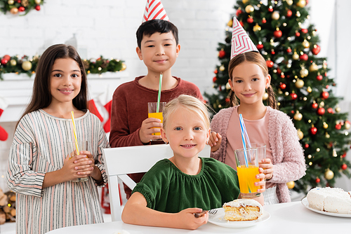 Smiling girl looking at camera near birthday cake and multiethnic friends with orange juice at home in winter