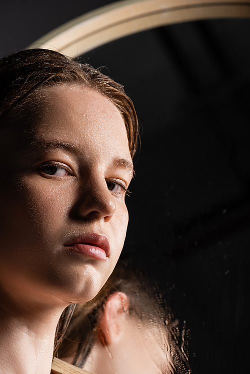 Young model with wet skin on face looking at camera near blurred mirror
