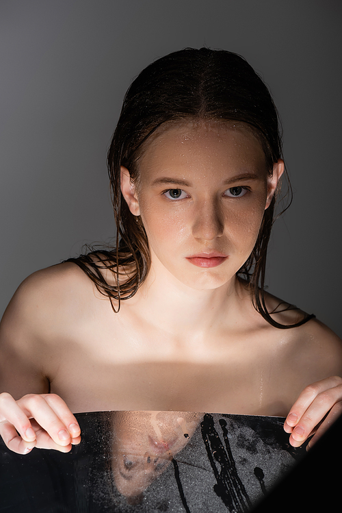 Young woman with wet skin and hair touching mirror isolated on grey