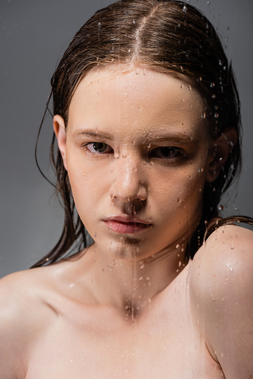 Portrait of woman with naked shoulders standing behind wet glass on grey background