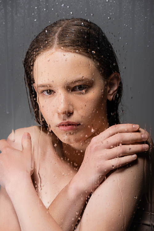 Portrait of young woman touching shoulders behind wet glass on grey background