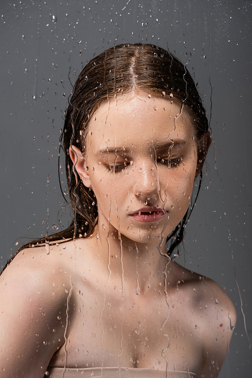Fair haired model with naked shoulders standing behind wet glass on grey background
