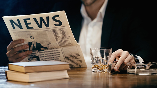 Cropped view of blurred businessman reading newspaper near whiskey and books on table isolated on black