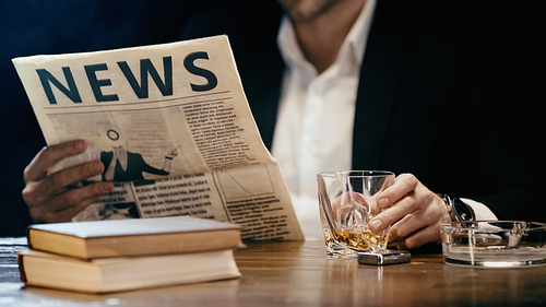 Cropped view of businessman reading newspaper and holding glass of whiskey near books isolated on black