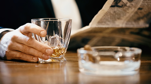 Cropped view of businessman holding glass of whiskey and newspaper near ashtray isolated on black