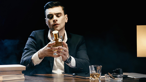 Businessman holding bottle of whiskey near books and cigar on table on black