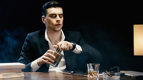 Businessman holding bottle of whiskey near books and ashtray on table on black